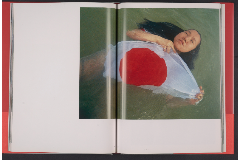 Artistic photograph of a woman floating in water and holding a Japanese flag. Her hair fans out around her, and her eyes are closed. 
