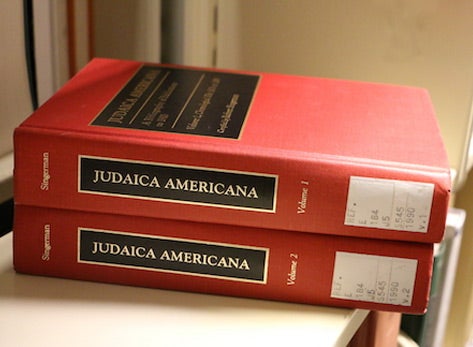 A photo of volumes 1 and 2 of Robert Singerman's JUDAICA AMERICANA stacked on a library shelf. The books have bright red ocvers and the title written in gold inside a black box.