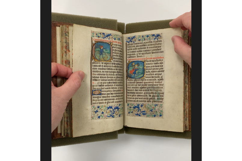 An illuminated manuscript being held open by two hands.