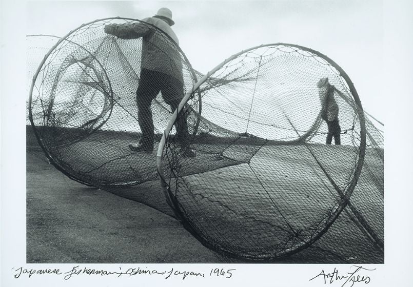 Black and white photograph shows two fishermen on the shore maneuvering large nets