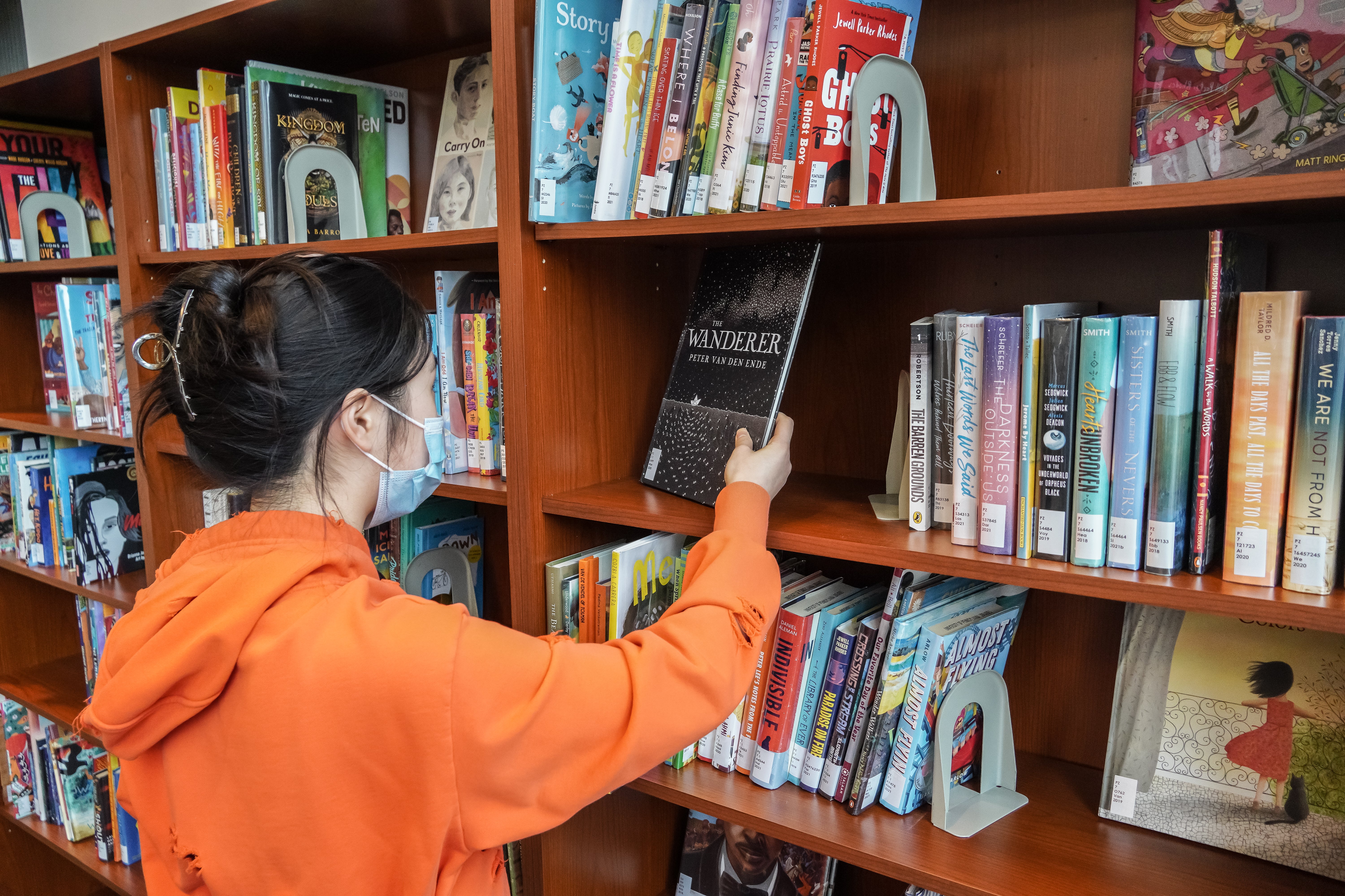 A person wearing a face mask stands in front of a bookshelf filled with colorful children's books.