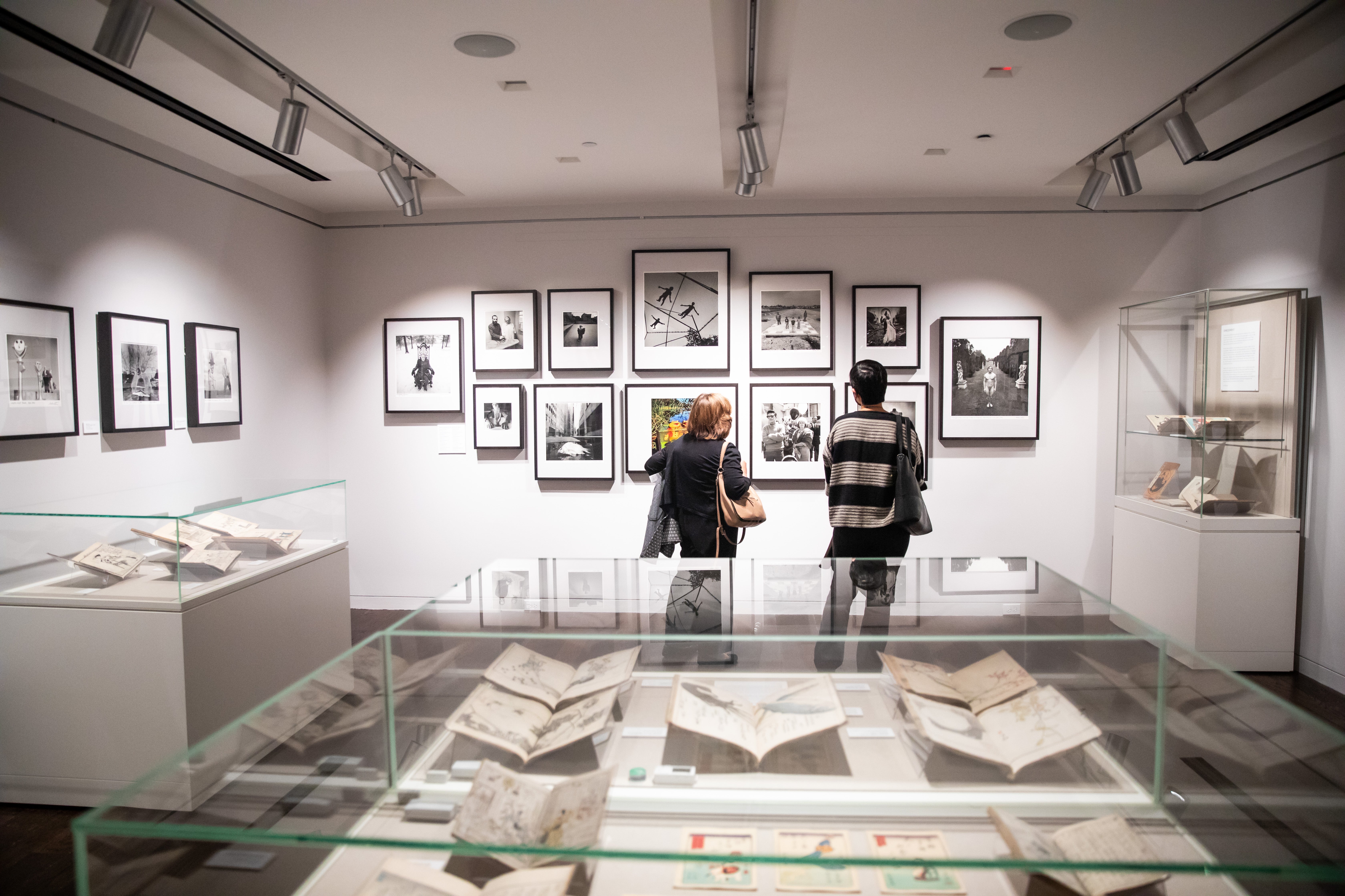 Inside an exhibit room, with a wall of 13 framed photos in the background. Two people with their backs to the viewer are looking at them. A glass exhibit case sits in the immediate foreground.
