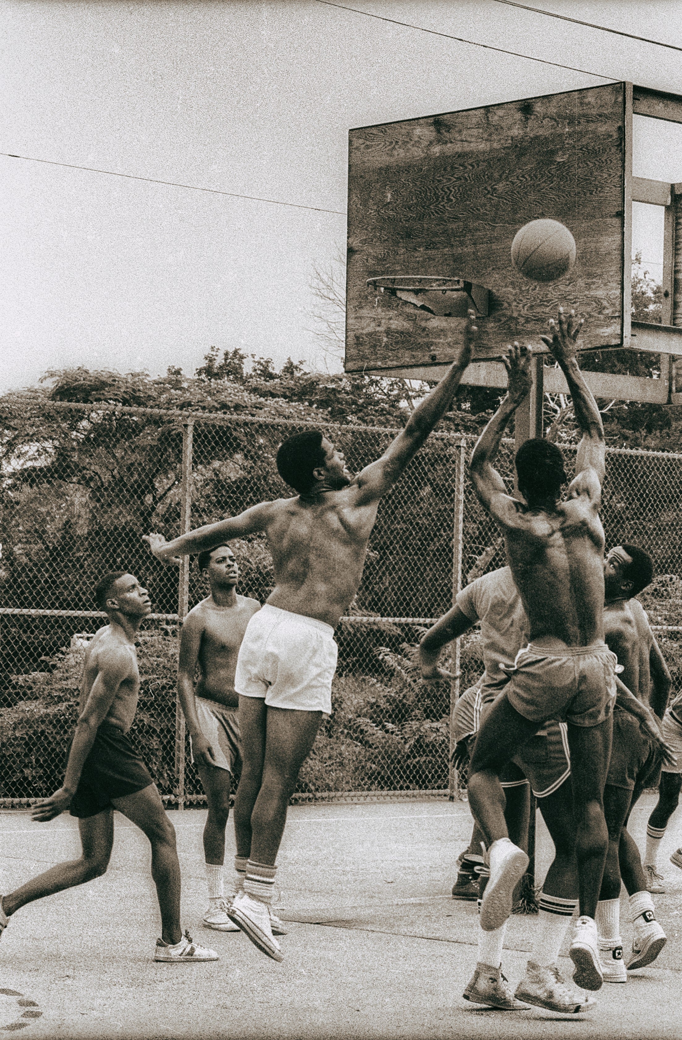 In a basketball game, a man is shooting the ball and a second man is leaping to block the shot while others look on.
