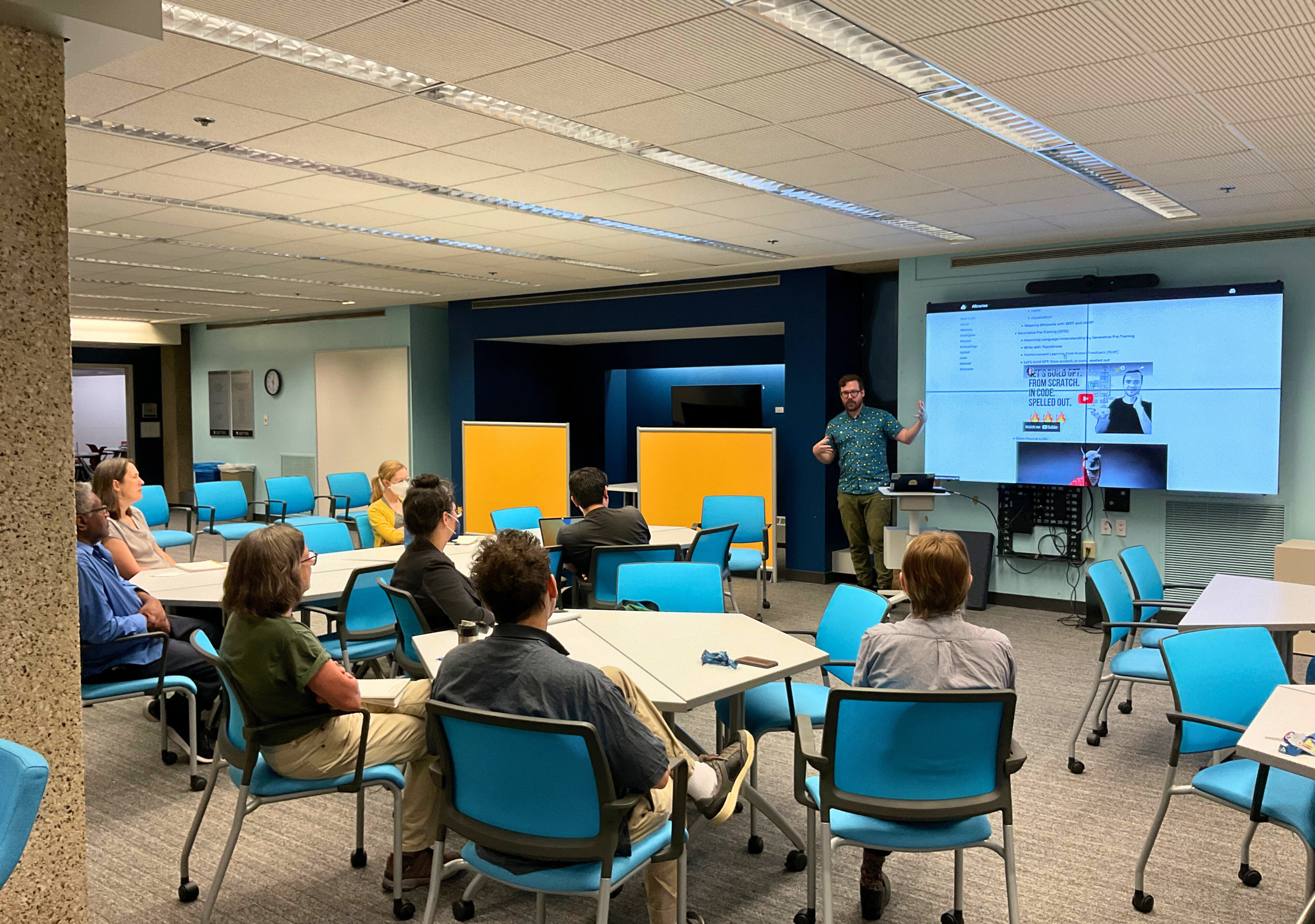 A large room filled with round tables and blue chairs. About a dozen people are sitting at the tables watching a person present from a projected powerpoint presenation.
