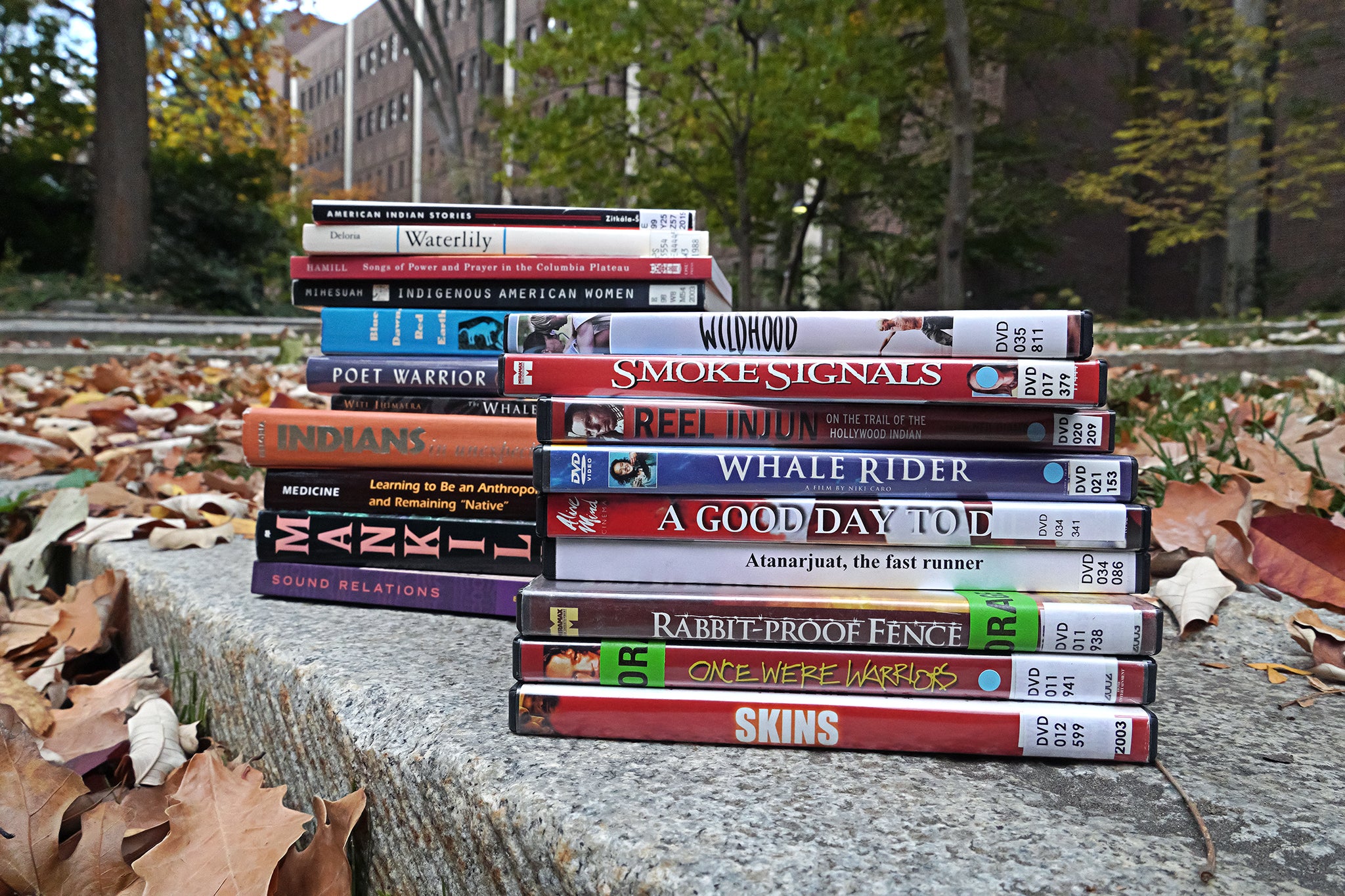 A multicolored stack of books sits next to a stack of DVDs on a rock surrounded by fallen leaves. A few of the titles include books American Indian Stories, Warrior Poet, and Sound Relations; and DVDs Smoke Signals, Rabbit-Proof Fence, and Skins.