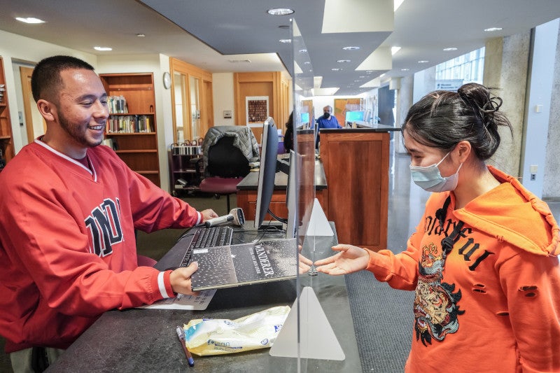 A student hands a book to a library assistant standing behind a desk.