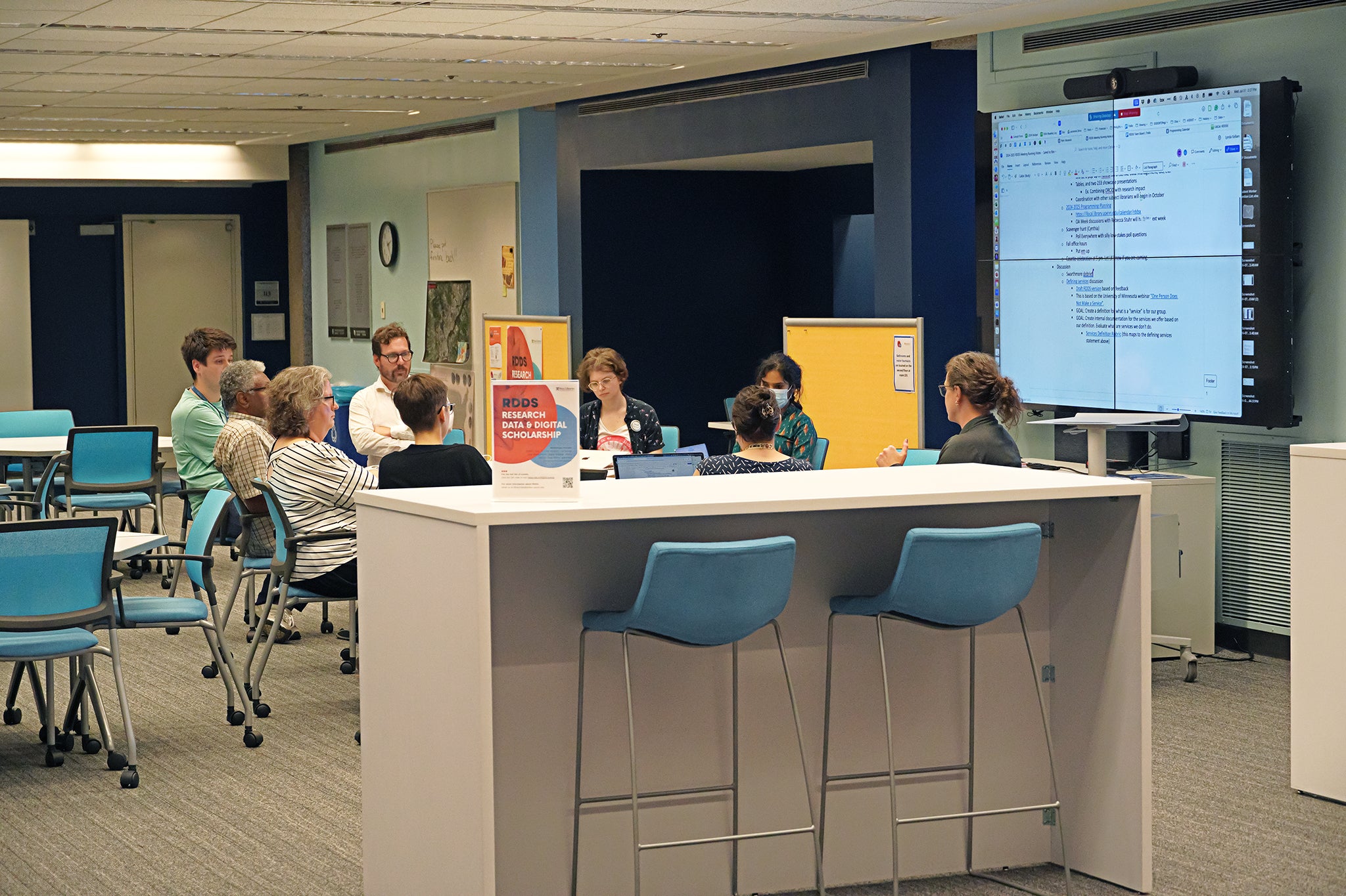 An indoor space shows a group of nine people seated in a semi-circle, facing a large screen on a wall. In front of and surrounding the group are tables and chairs of different heights. Posters on the wall, including one promoting “Design Thinking,” suggest a focus on creativity and innovation. Participants have laptops and notepads, indicating active involvement in the session.