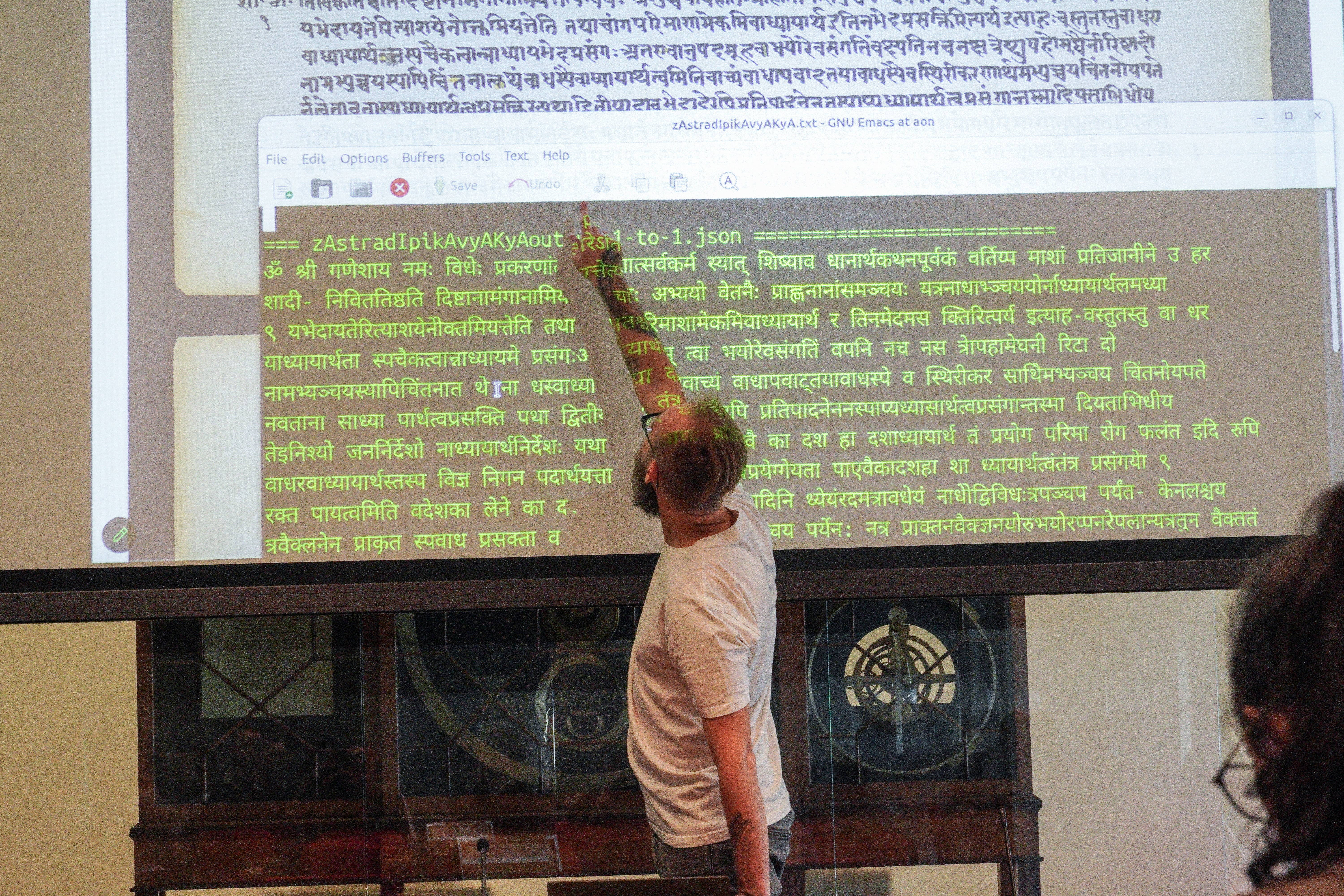 man pointing to a projection with two windows. The window at the back is the image of a Devanagiri script manuscript. The window in front is a black with green machine readable text of the Devanagari text.