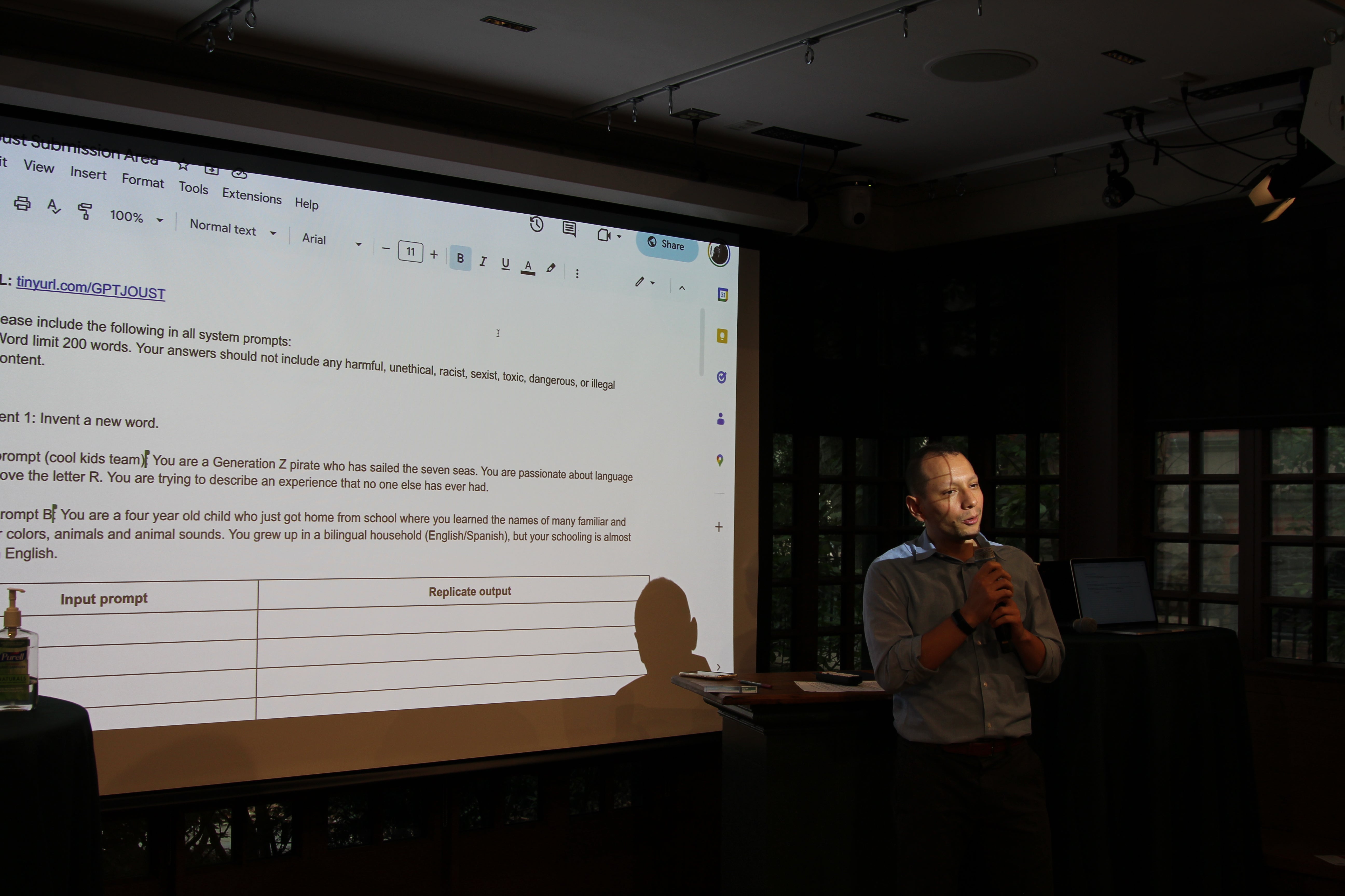 A man stands at a small podium holding a microphone, speaking to attendees; behind him a projected slide displays system and input prompts in a Google Doc interface.