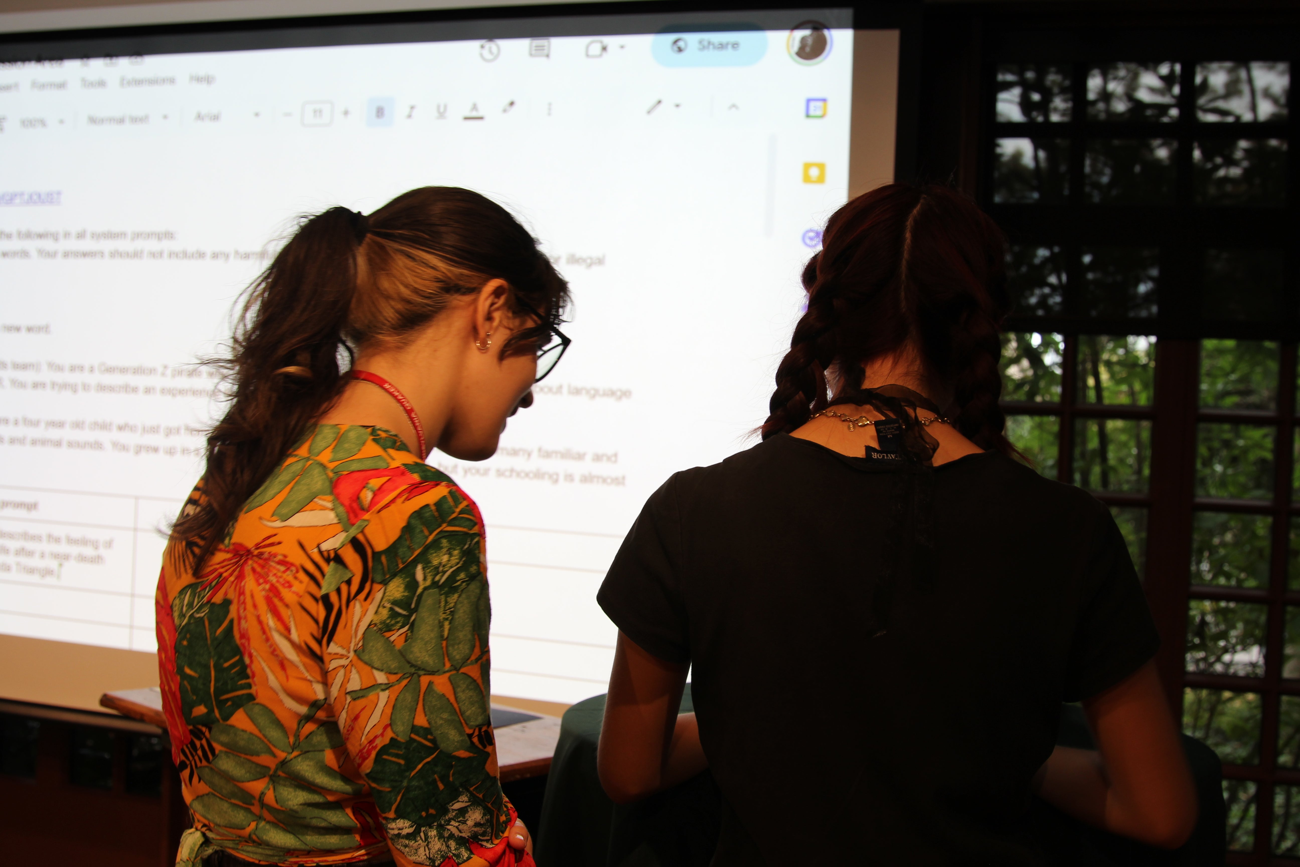 Two participants stand at the front of the room entering their custom prompts into the live Google Doc interface projected on the screen, collaborating quietly before the next round begins.
