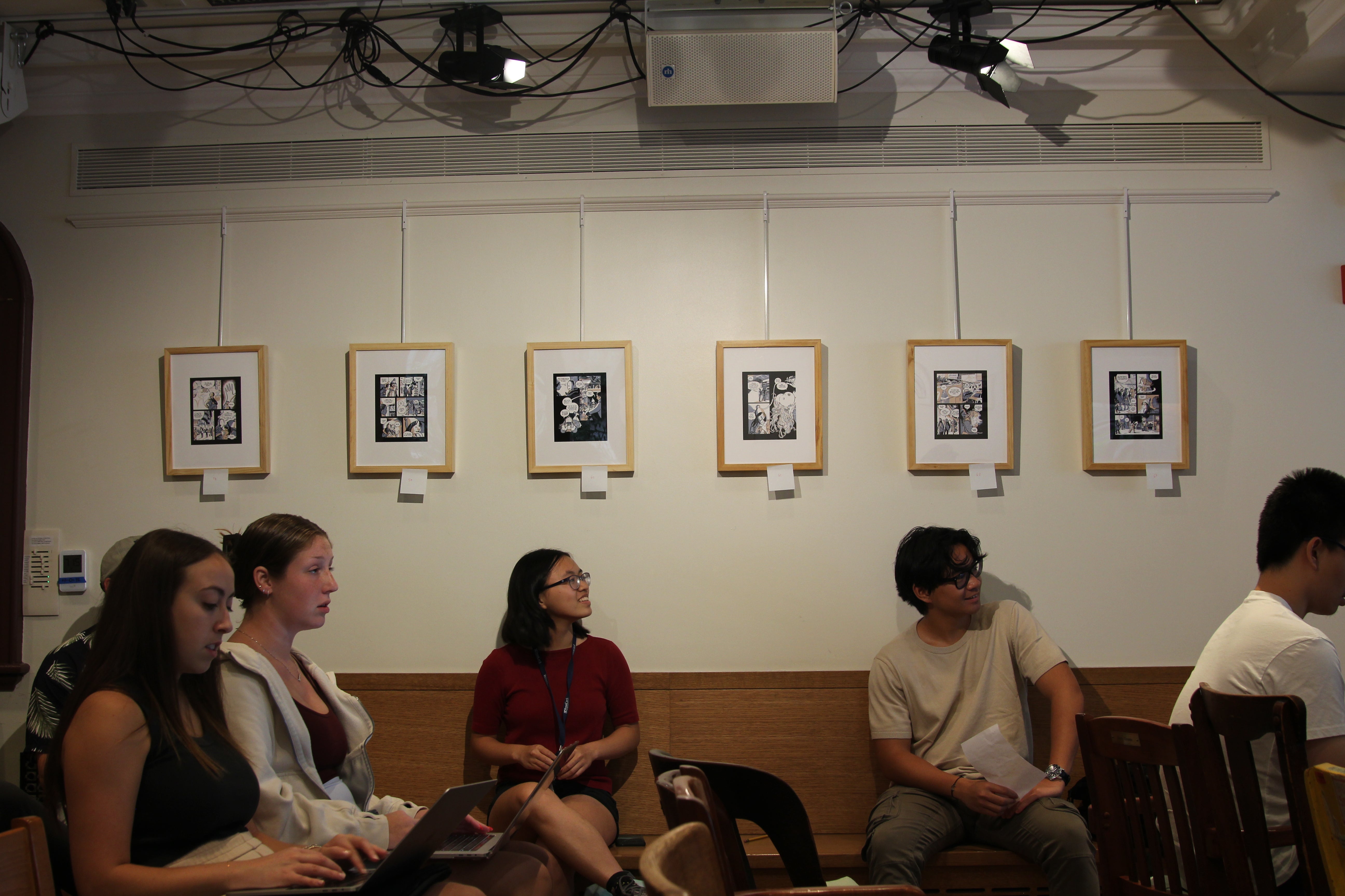 A row of framed black-and-white comic panels hangs on a light-colored wall above seated workshop participants; some have laptops on their laps, all looking toward the front of the room.