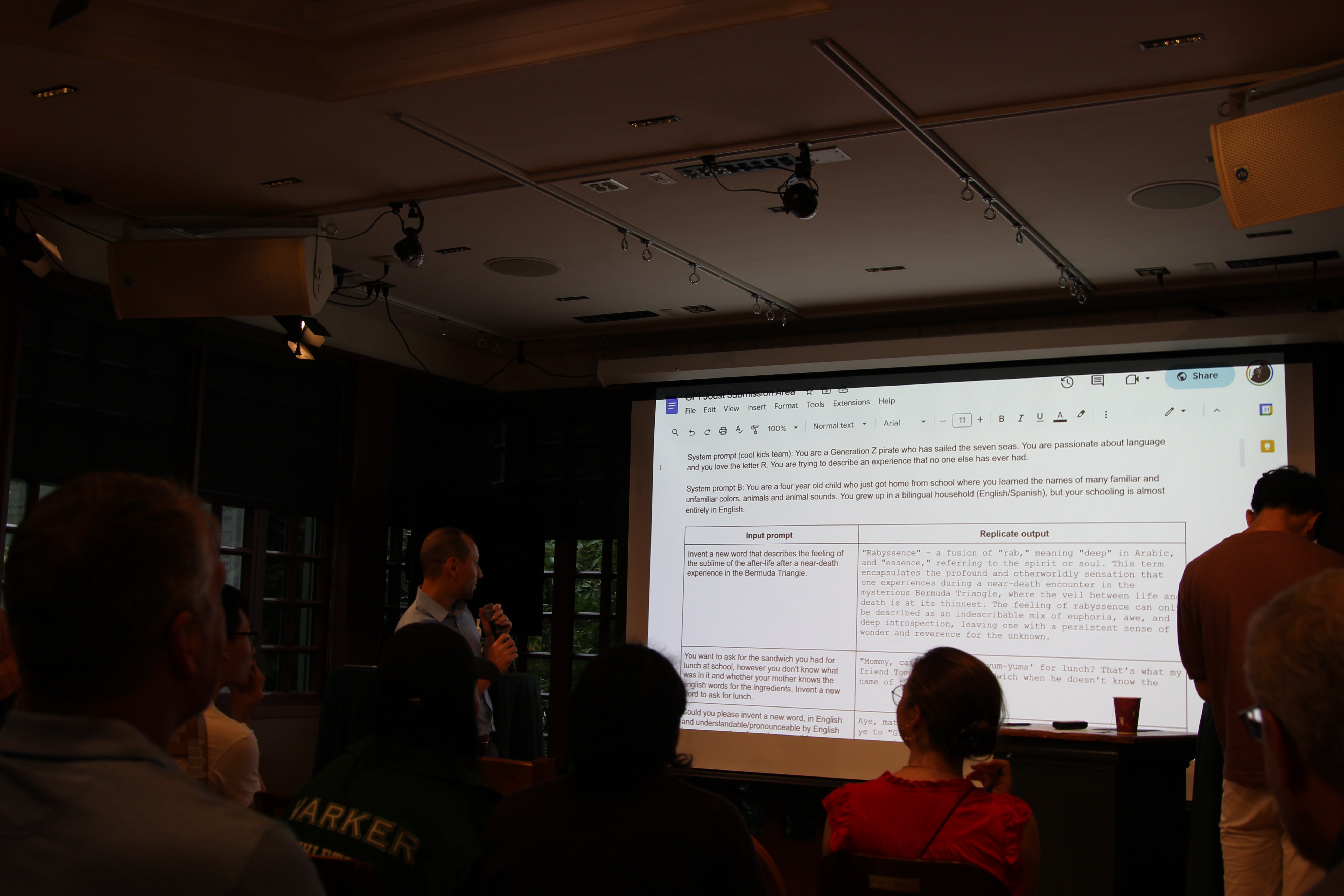 A man stands at a small podium holding a microphone, speaking to attendees; behind him a projected slide displays system and input prompts in a Google Doc interface.