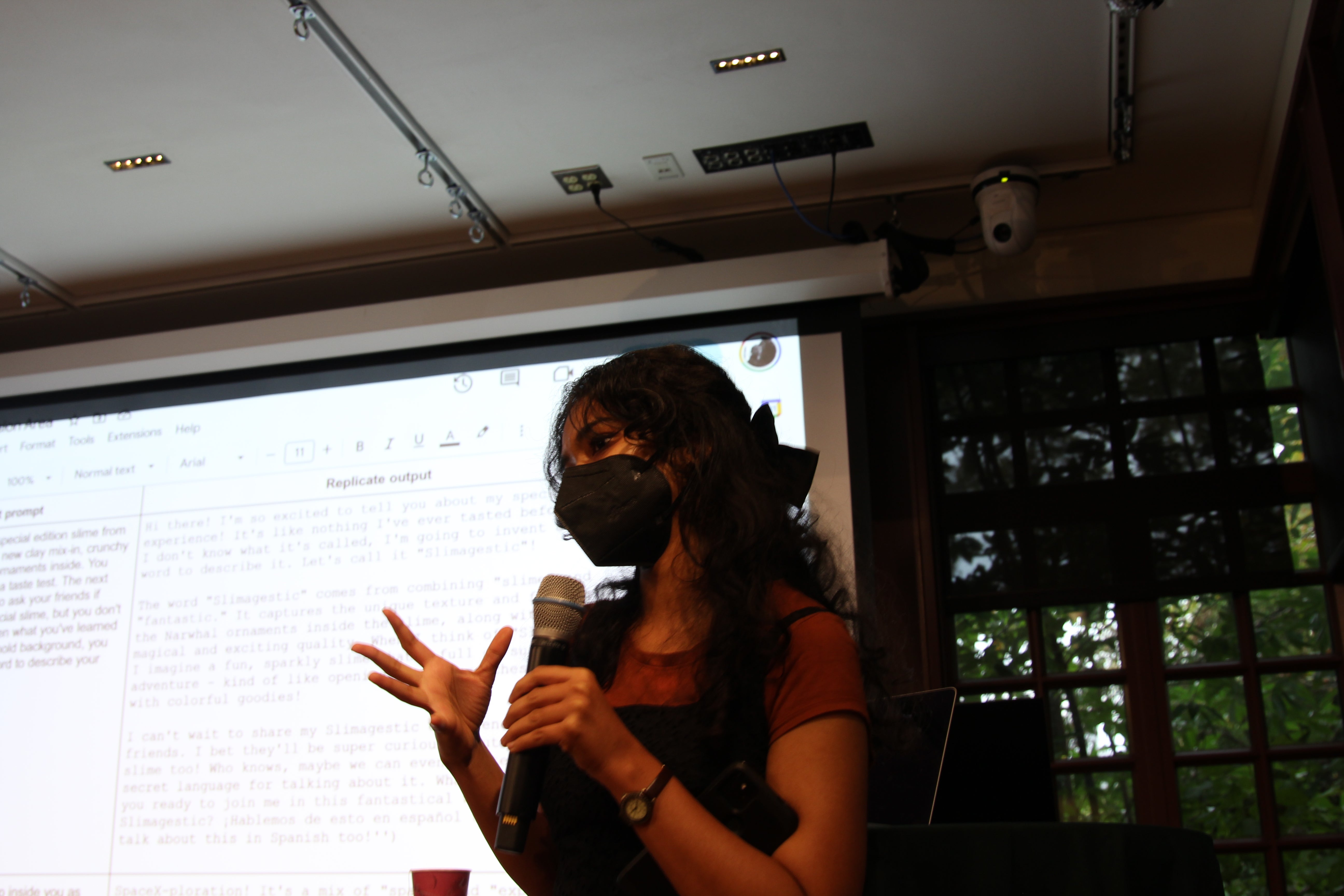 A young woman wearing a black face mask and holding a microphone gestures with her hand as she speaks; behind her, a projected slide displays AI-generated text in a side-by-side comparison format.