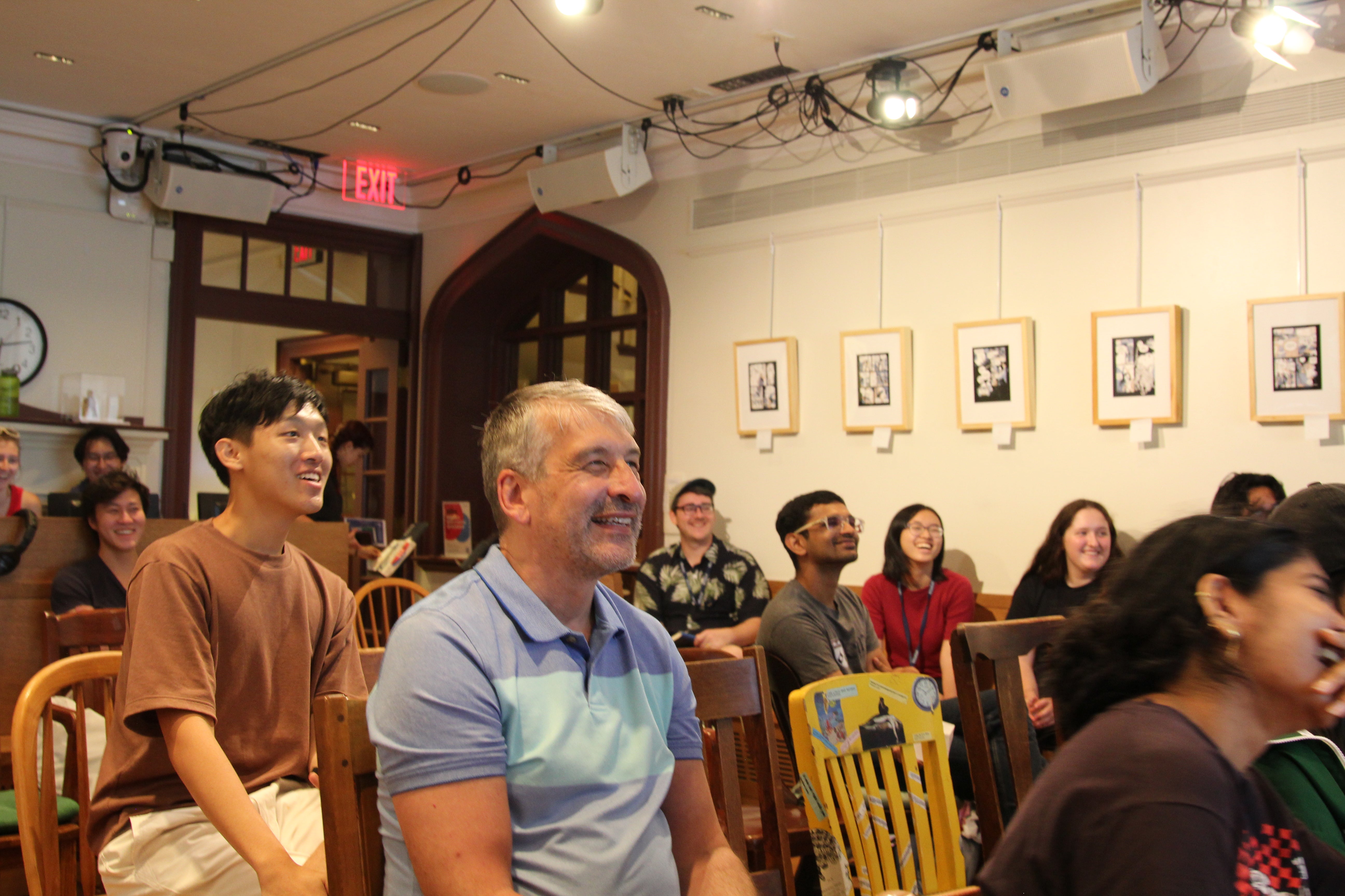 Workshop participants standing and seated in a room with framed artwork on the walls, all looking toward the front with amused expressions as winners are declared.