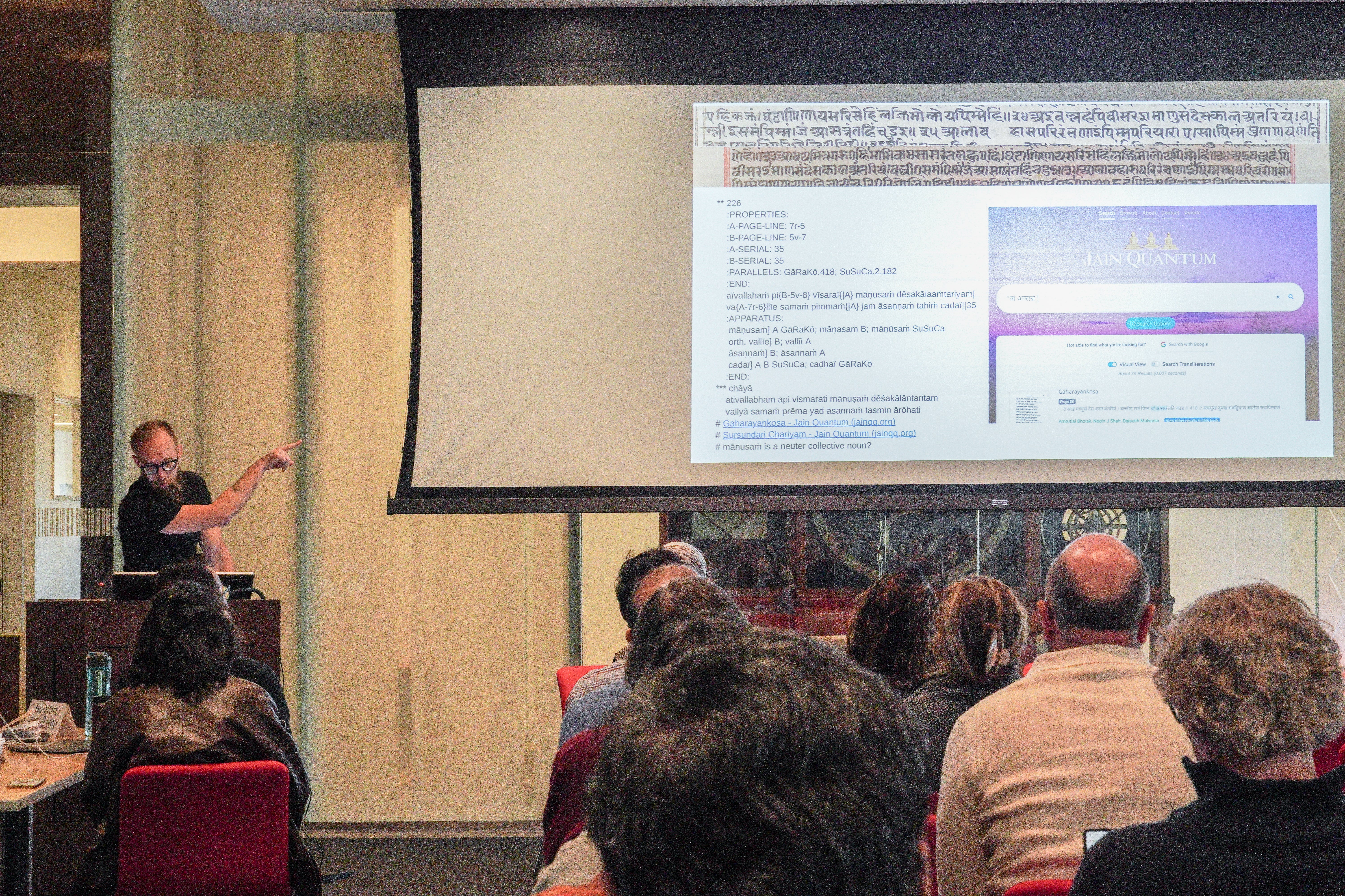 man pointing to screen from a podium. the screen has sanskrit script text