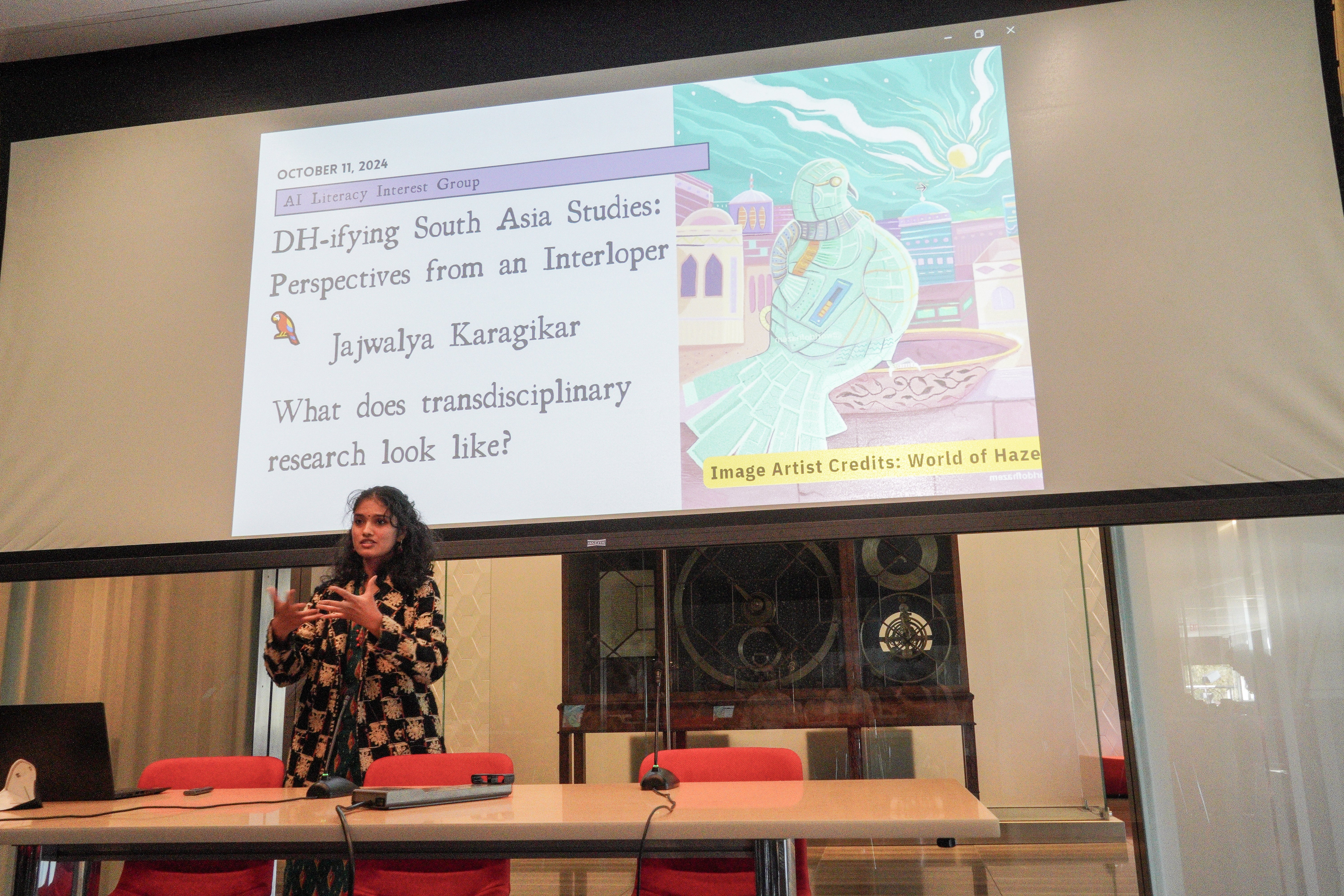 woman wearing beige and black knitted coat in front of a slide with parrot and words "DH-ifying South Asian Studies: Perspectives from an Interloper"
