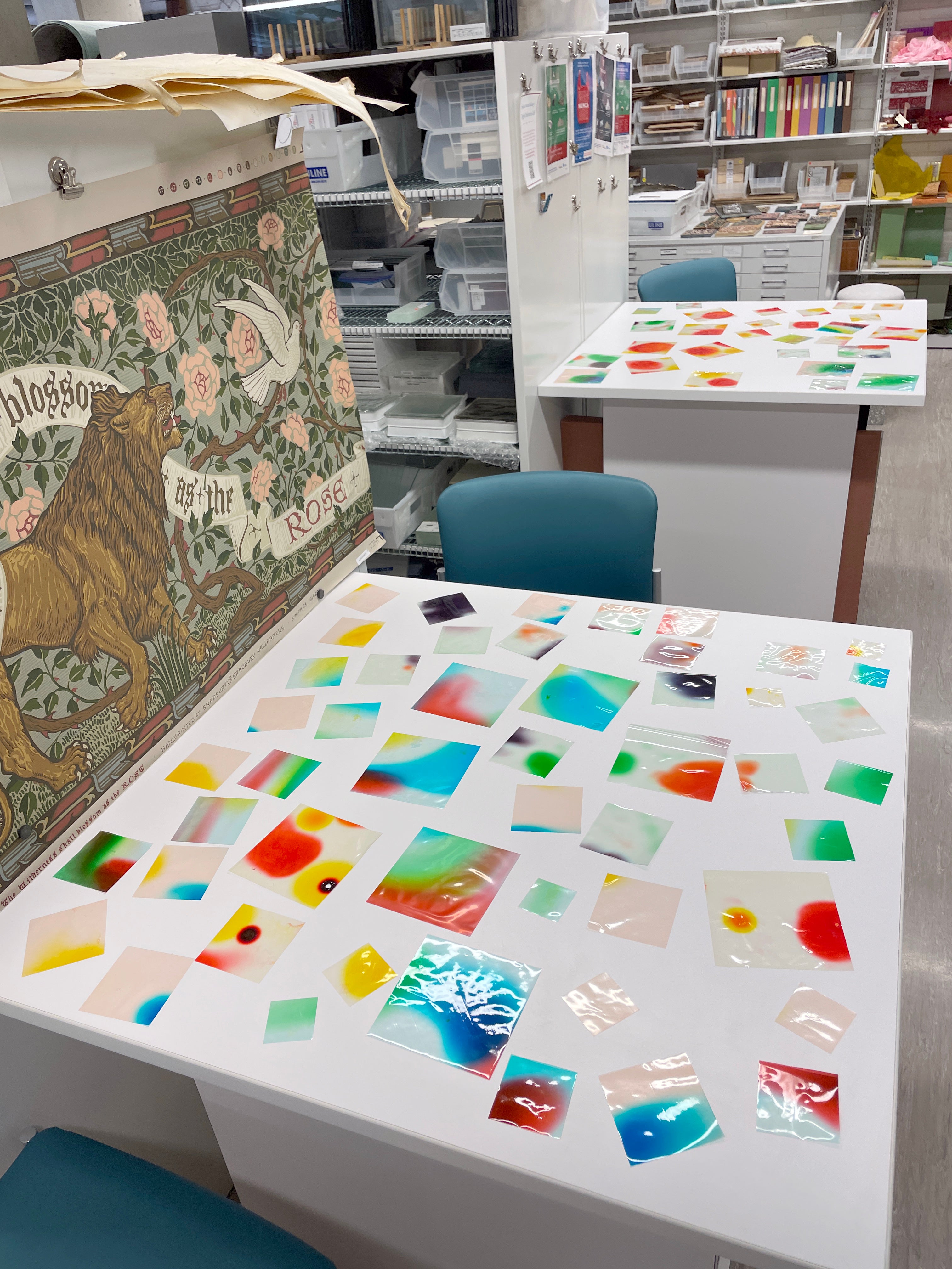  Two white tables covered with various colorful, translucent bioplastic sheets. One table also has a large framed print of a floral and animal design.