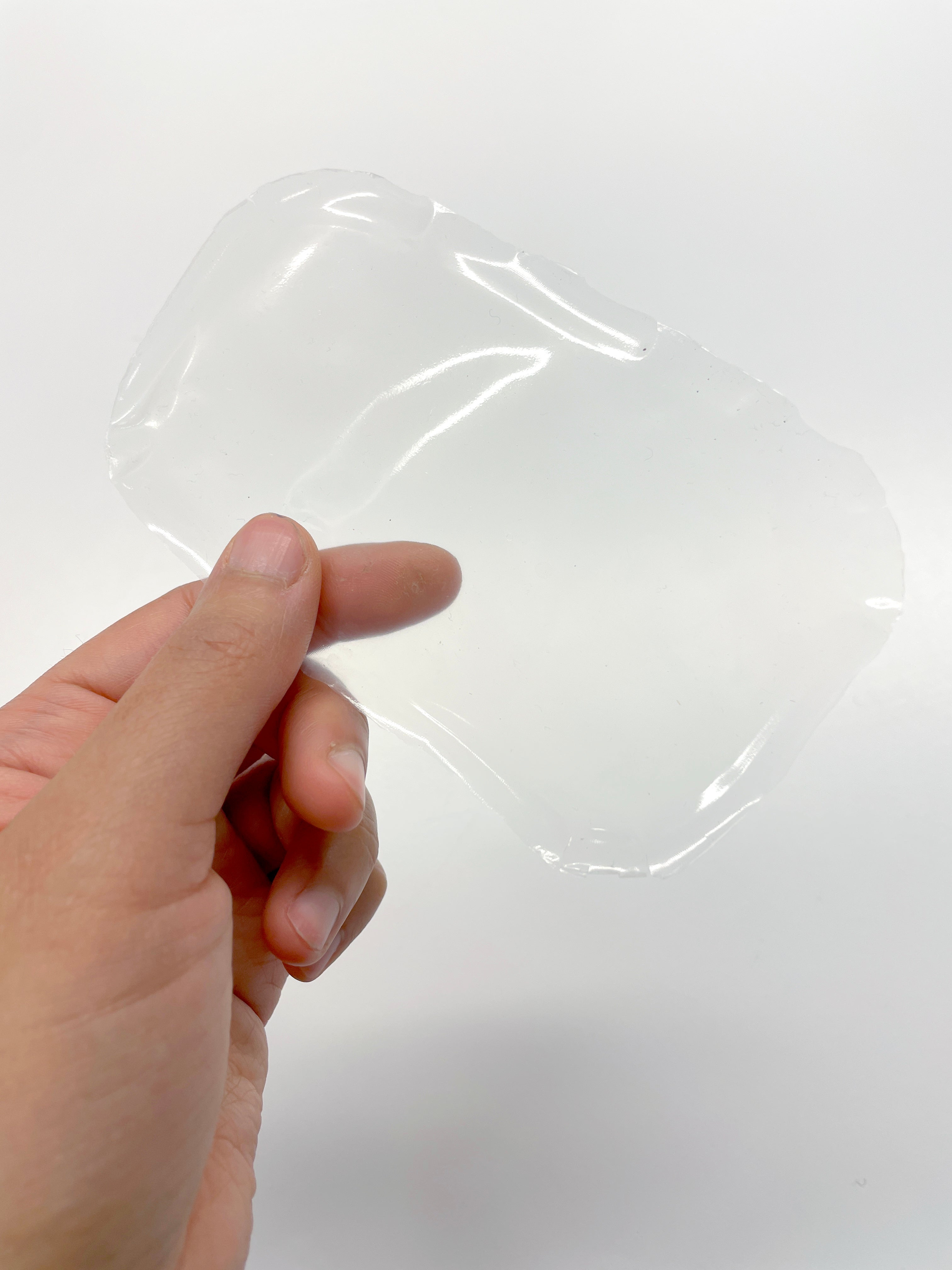 A hand holds up a thin, translucent, and slightly irregular sheet of gelatin, showcasing its flexible and somewhat reflective surface against a white background.