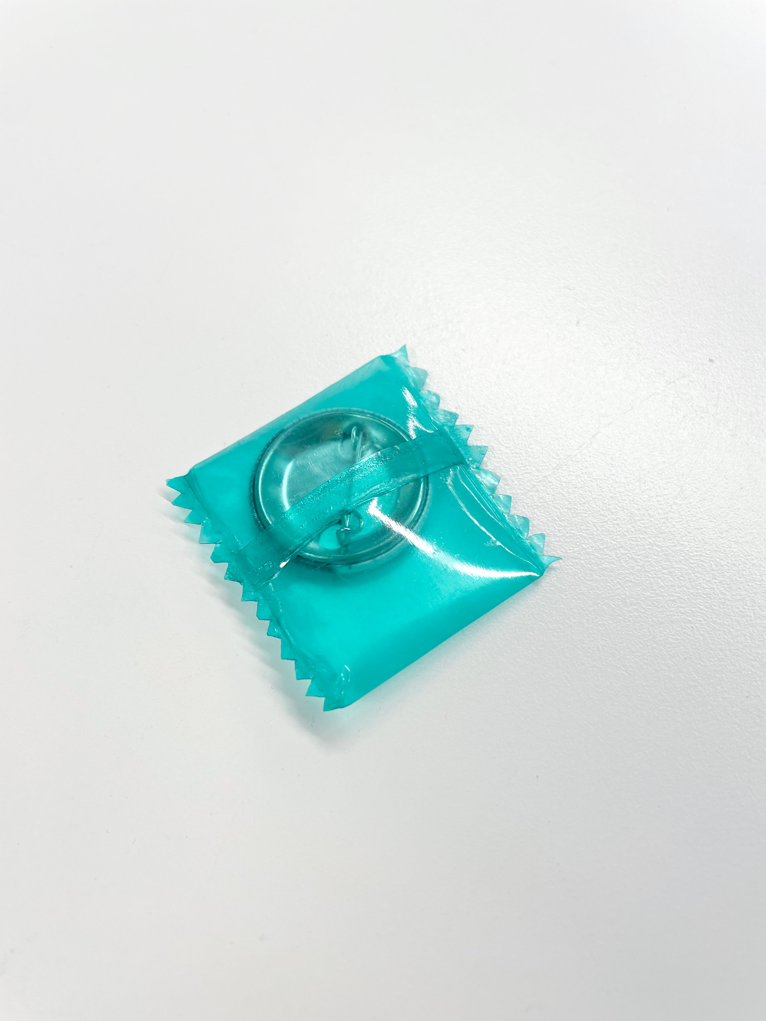  A clear, teal-tinted plastic wrapper with zigzag edges encloses a circular, metallic object. A matching teal ribbon is visible around the object.