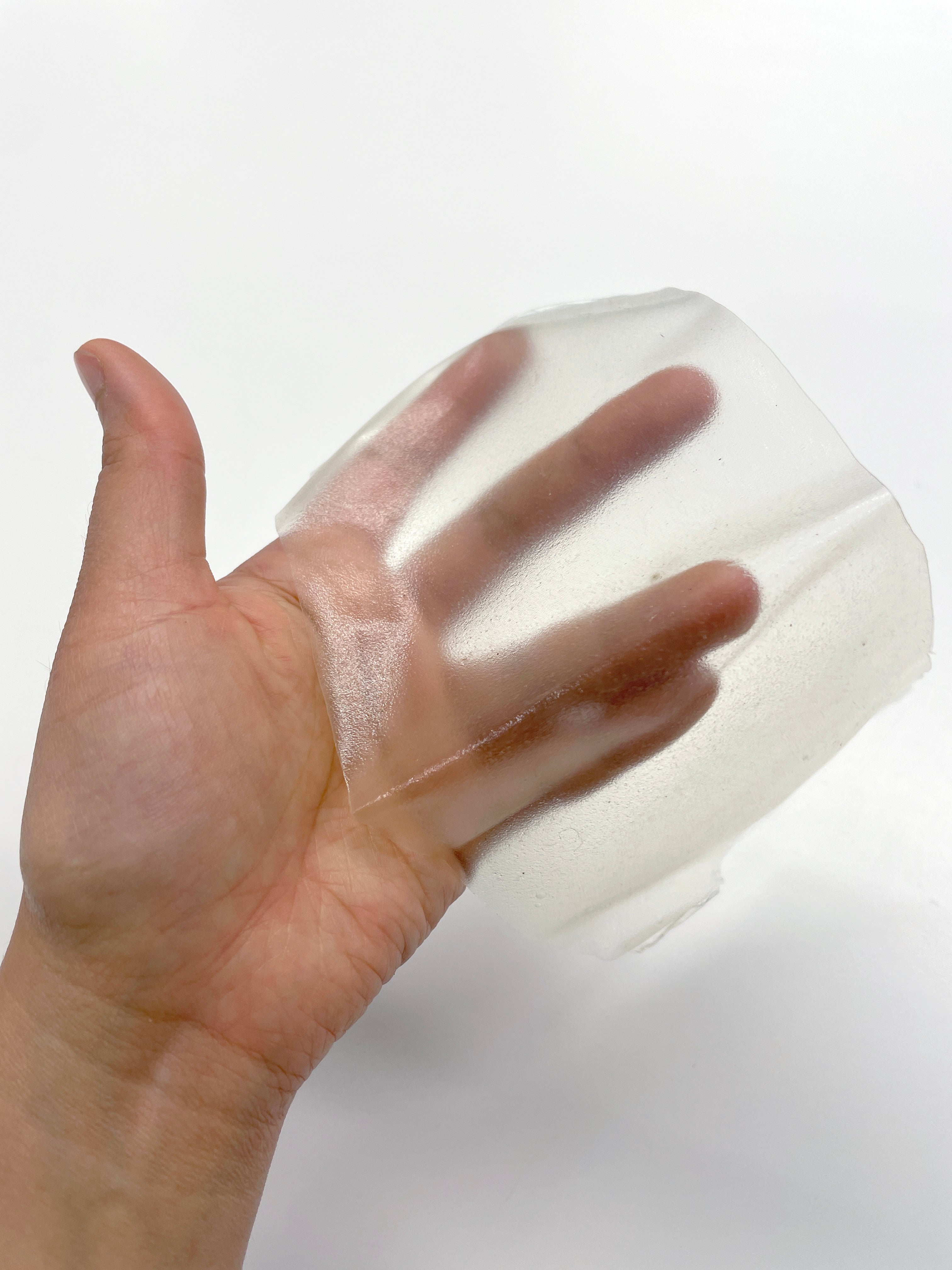  A hand holds up a thin, semi-translucent, and slightly irregular sheet of agar bioplastic, showcasing its flexible and somewhat reflective surface.