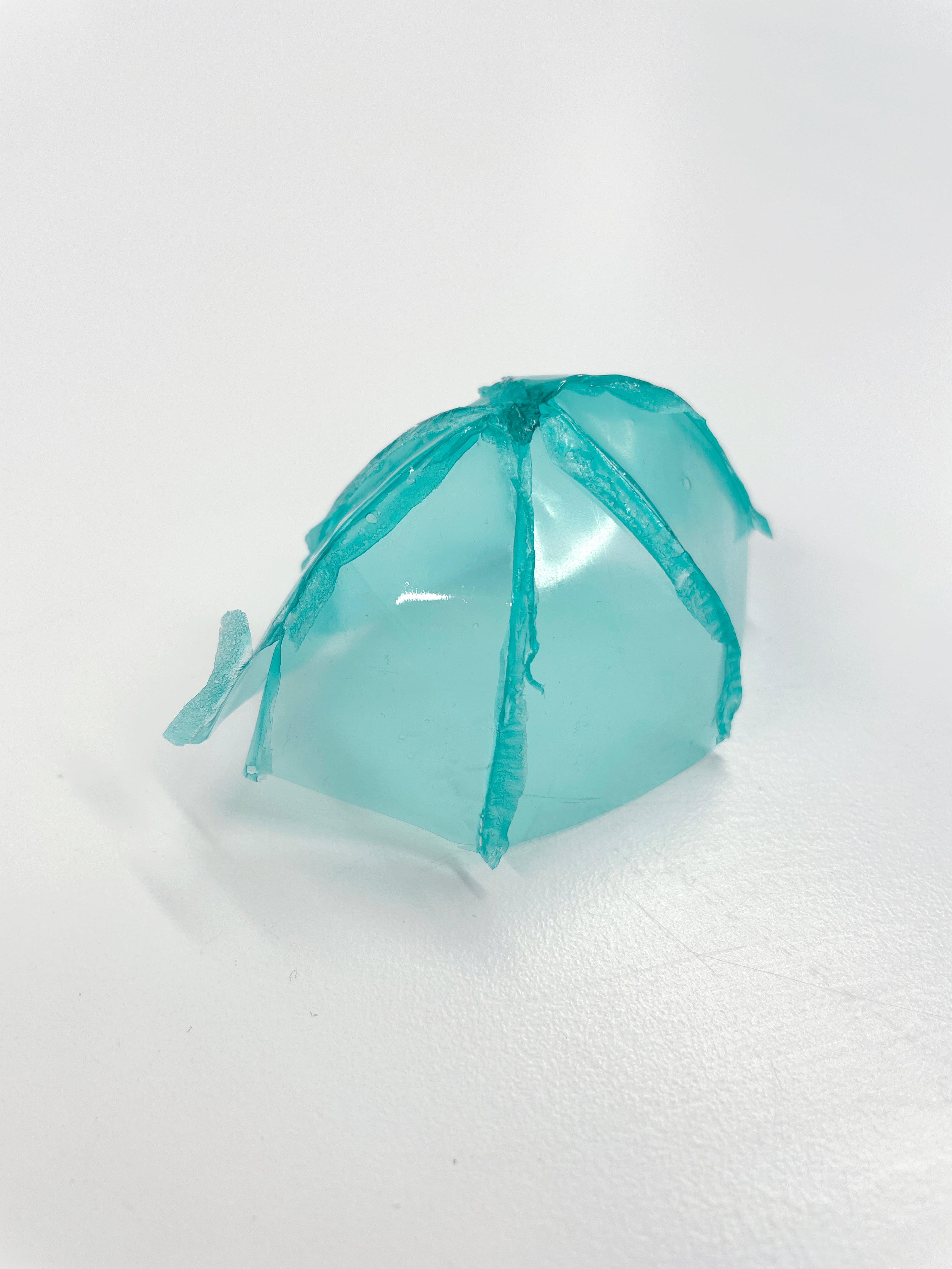  A transparent, teal, dome-shaped object with visible seams and a slightly irregular base, sitting on a white surface.