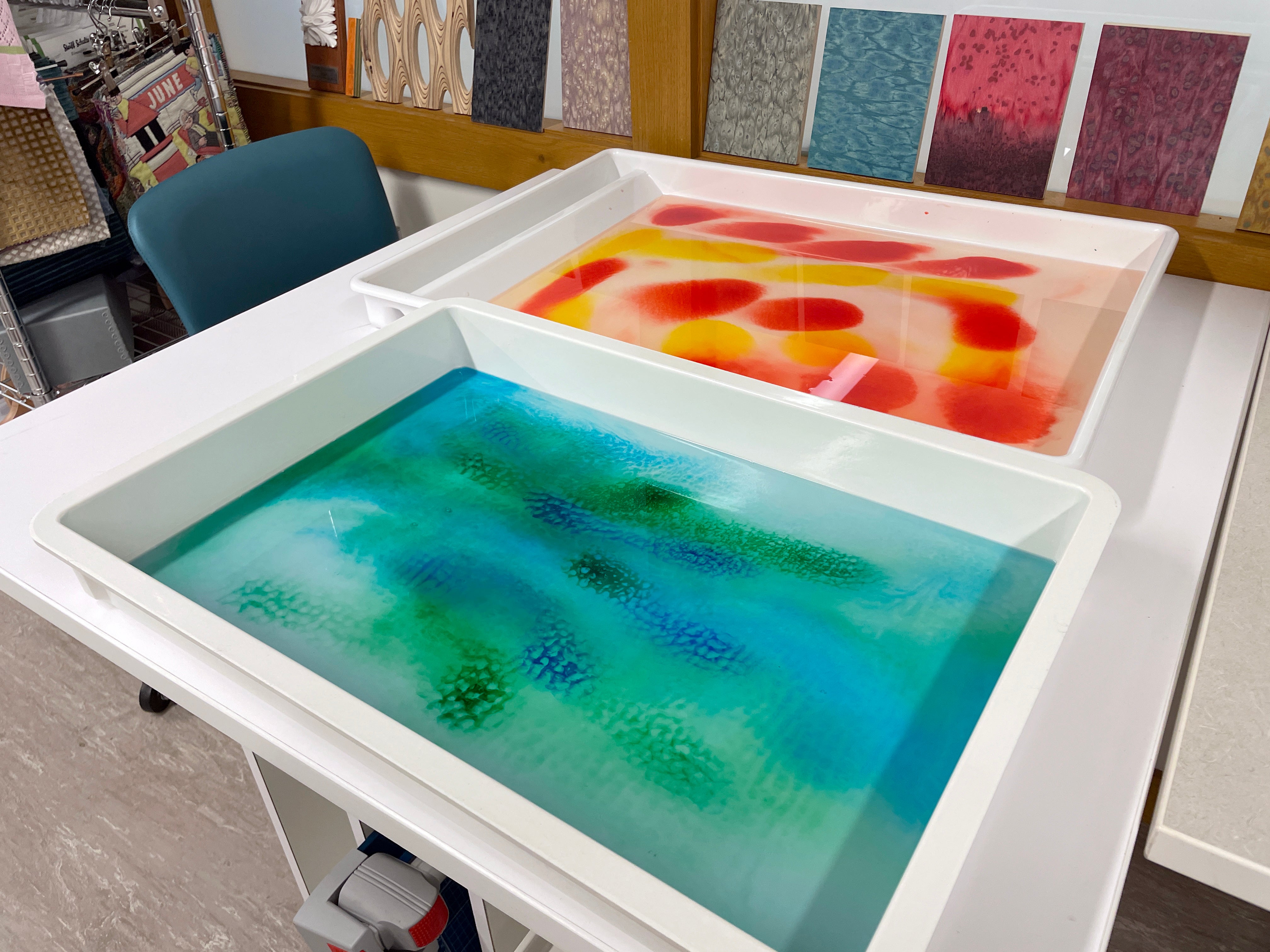 Two white trays on a table. The front tray holds a blue-green liquid, and the back tray contains yellow and red liquids.