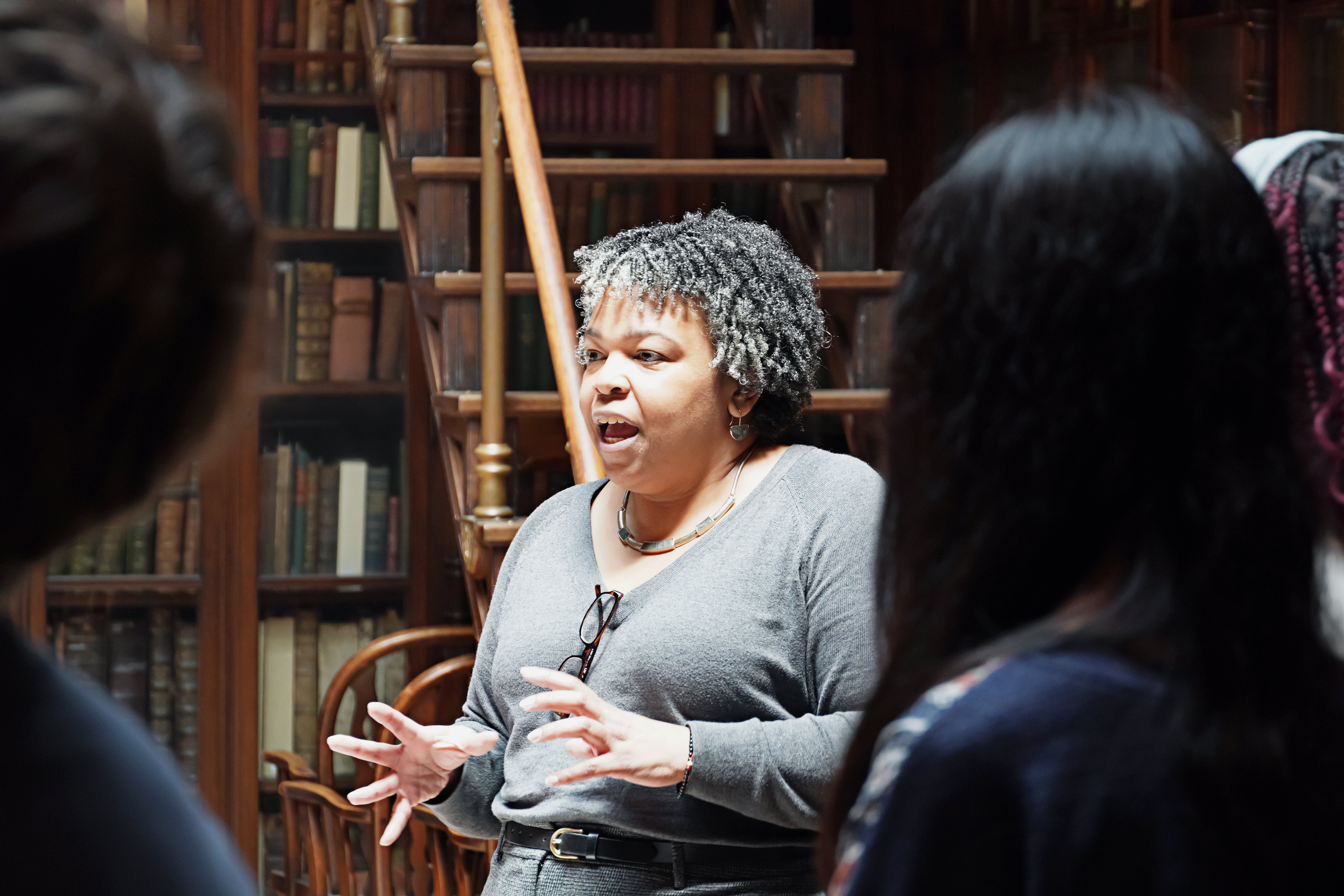 A woman in a library is speaking animatedly while gesturing with her hands. She is surrounded by books and a wooden staircase.