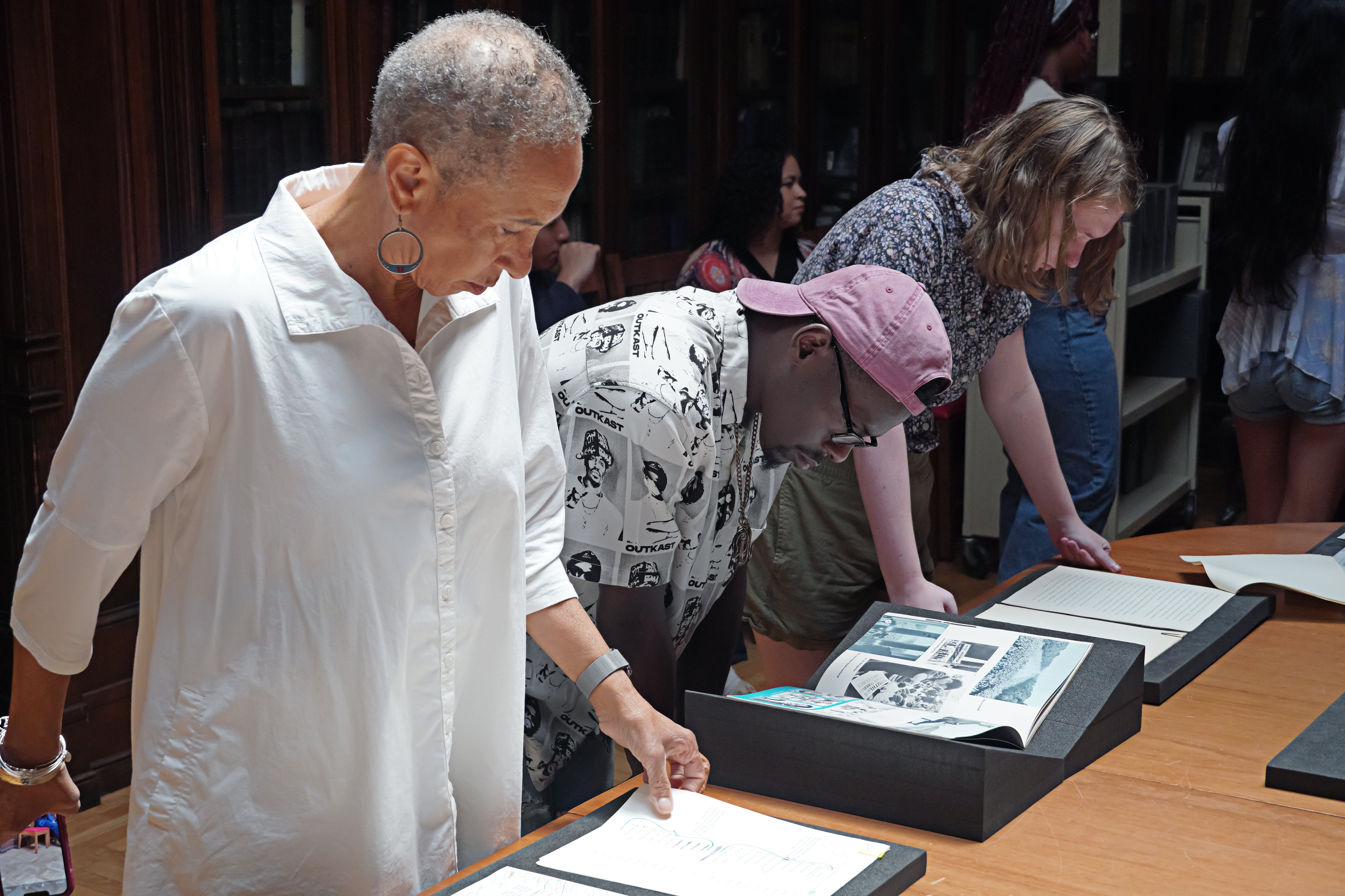 A small group of students and an instructor look down at documents displayed on a wooden table inside a library.