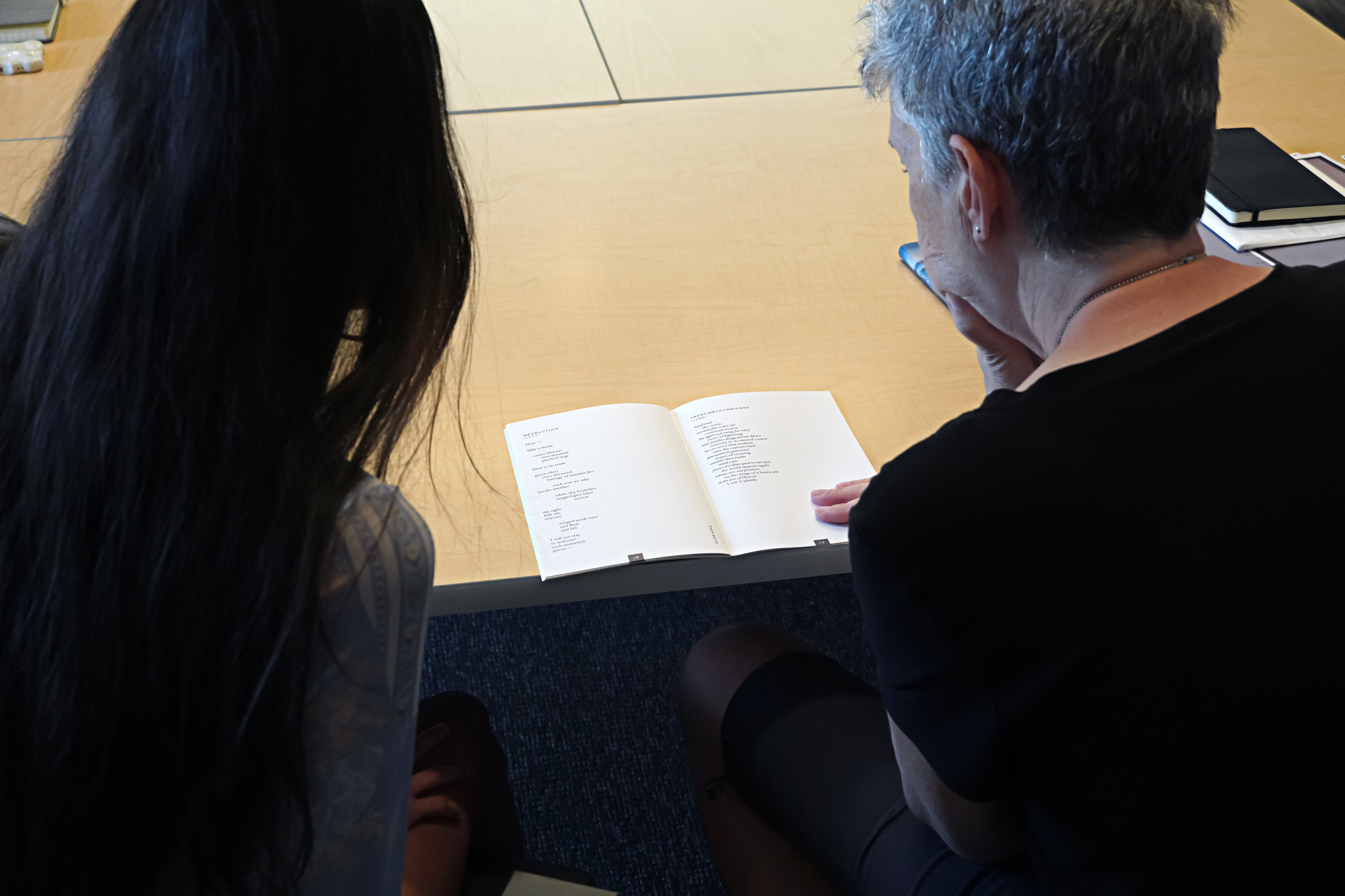 Two people, one with long dark hair and one with short gray hair, lean over a table and read from a book of poetry.