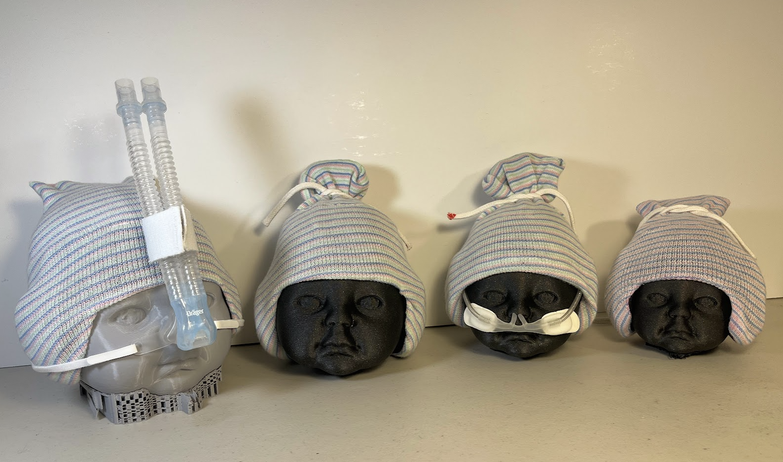 Four 3D-printed infant head models wearing striped neonatal beanies in different sizes, with some models fitted with nasal cannulas or CPAP tubes to demonstrate compatibility with respiratory support equipment