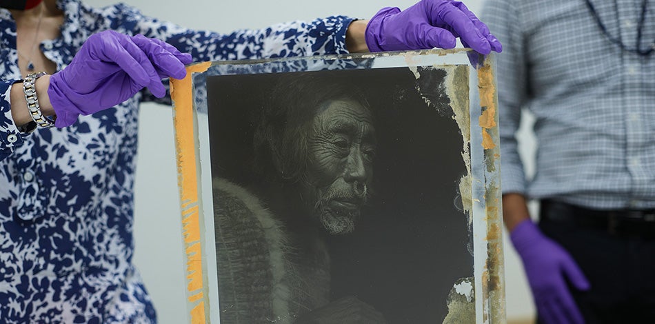 Individual with gloved hands holds up a glass plate showing a black and white photographic image of a Native American man.