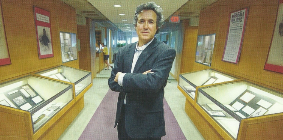 A man stands with his arms crossed in a hallway lined with glass cases of documents.