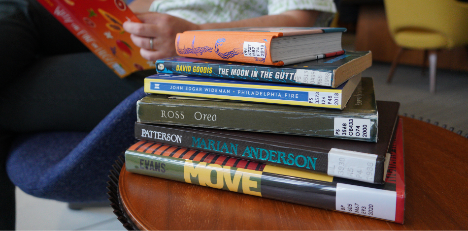 A stack of books on a side table in the library. A person is reading in the background.
