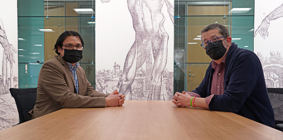 Two people with face masks sit at a meeting table and look to the viewer. An anatomical illustration adorns the wall.