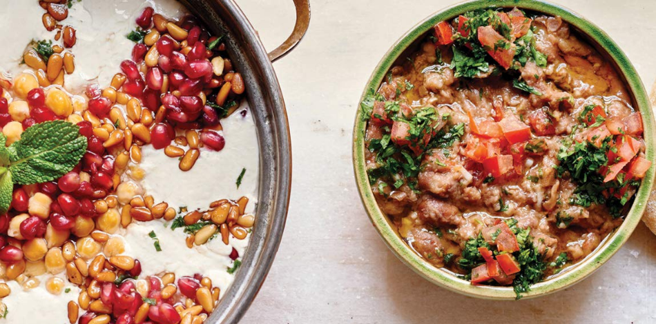 Two bowls of food: the one on the right is a bubbling curry-like dish topped with brightly-colored tomato and parsley; the one on the left is a creamy white soup topped with chickpeas, pomegranate, and pine nuts.