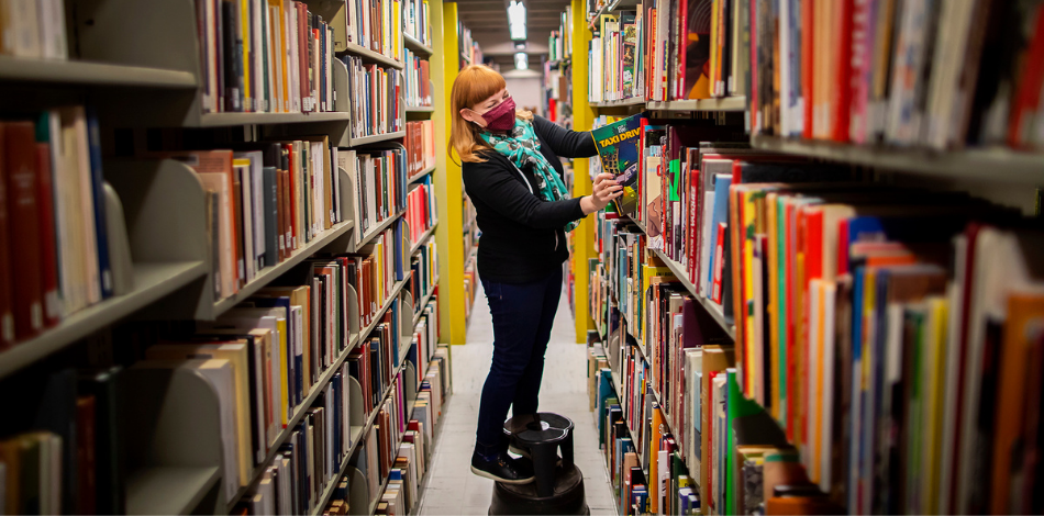 A woman stands on a round stool between two tall bookshelves.