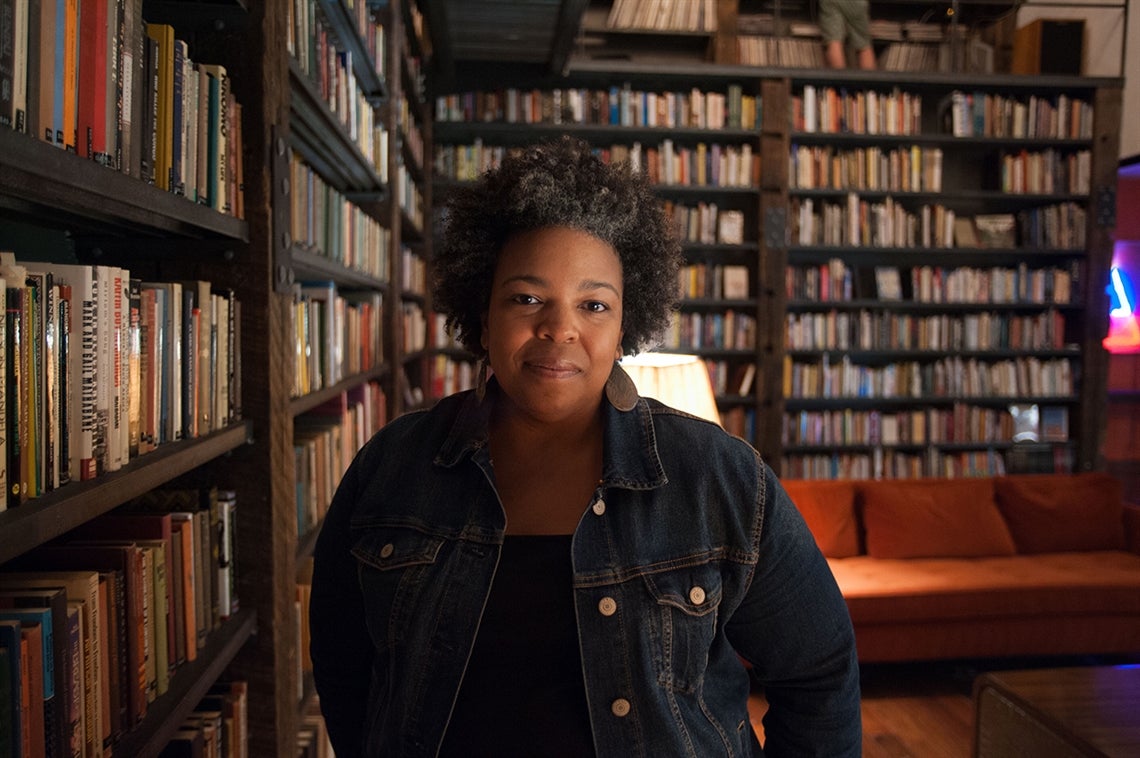 A woman stands in a softly-lit library surrounded by bookshelves.