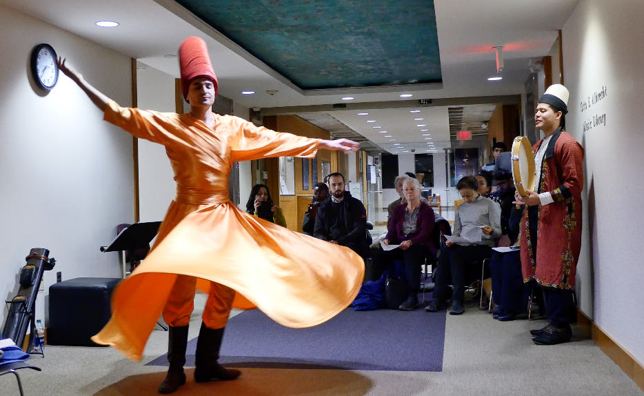 A dancer and a drummer perform indoors for a small crowd.