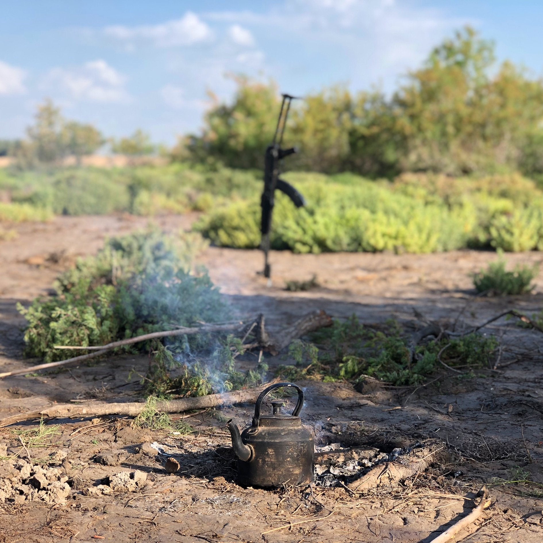 A steaming pot of tea on the ground. Out of focus behind it an assault rifle is propped up.